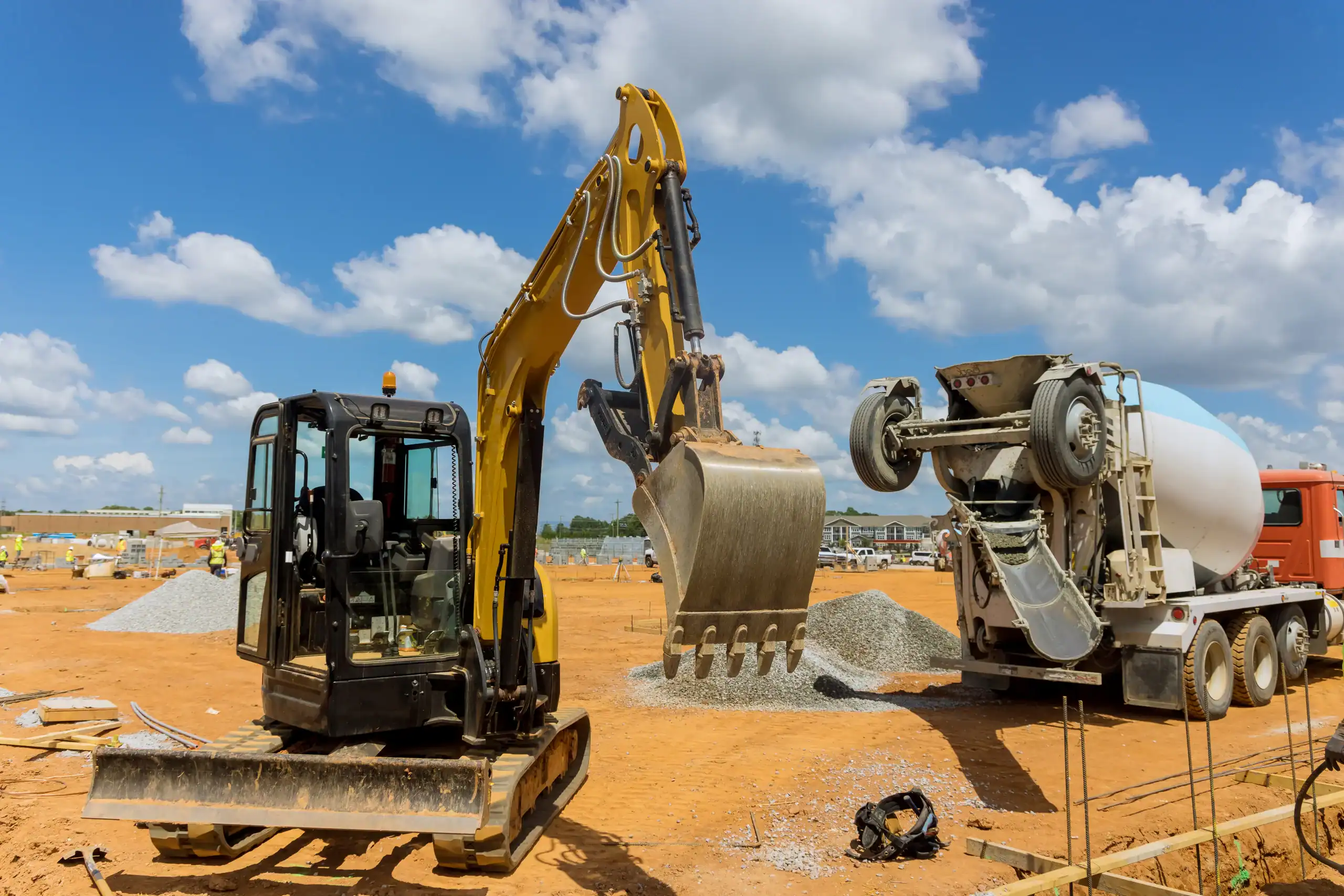 A yellow excavator, available through equipment rental St. Lucie & Orange County, FL, loads gravel beside a cement mixer truck at a construction site on a sunny day with blue skies and scattered clouds in the background.