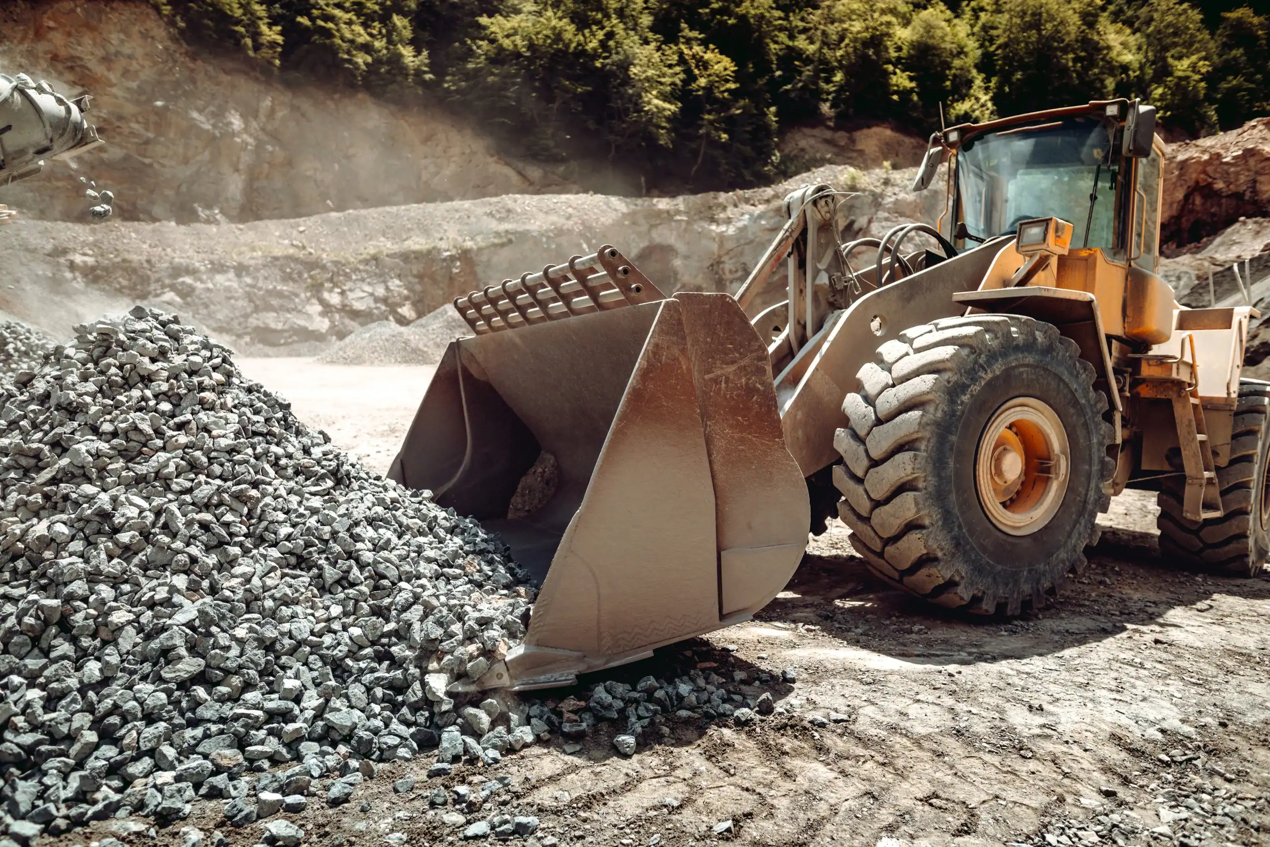 A large yellow front-end loader, available through equipment rental St. Lucie & Orange County, FL, moves a pile of gray rocks at a quarry, with trees and rocky hills in the background under bright sunlight.
