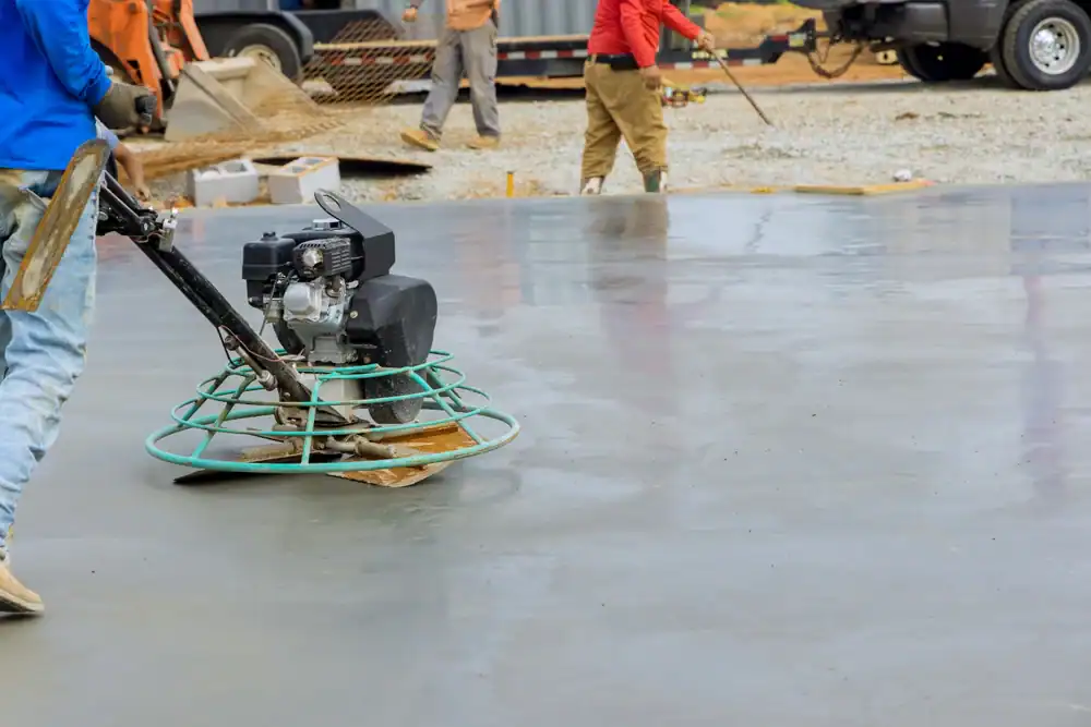 A construction worker uses a power trowel, available through equipment rental St. Lucie & Orange County, FL, to smooth and finish a large concrete surface at a building site while another worker stands in the background.