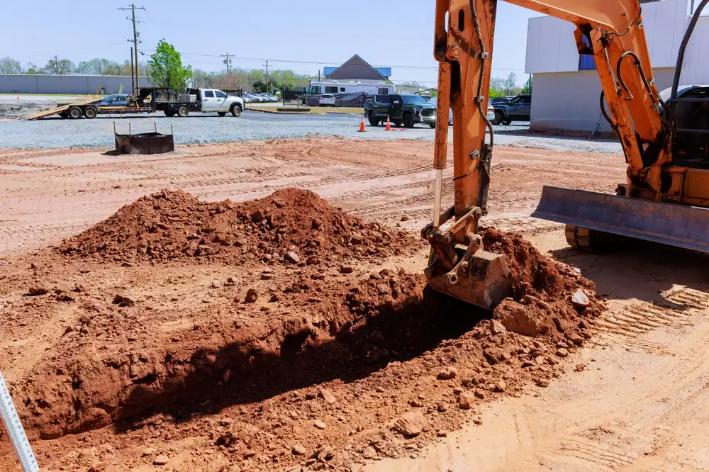 A construction site in FL features an excavator from equipment rental St. Lucie & Orange County digging a trench in reddish soil. Dirt piles and vehicles are visible in the background under a clear sky.