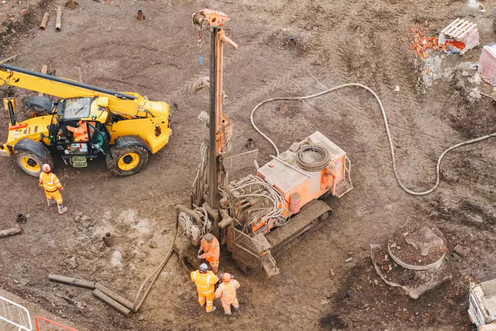 Aerial view of three construction workers in orange safety gear operating heavy drilling machinery—available through equipment rental St. Lucie & Orange County, FL—on a muddy site, with a yellow vehicle parked nearby.