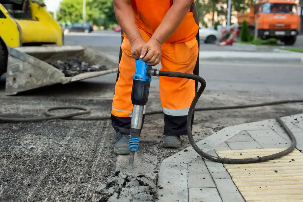 A construction worker in orange safety clothing uses a jackhammer to break up asphalt on a road near a sidewalk curb in FL. Construction equipment and traffic, possibly from equipment rental St. Lucie & Orange County, are visible in the background.