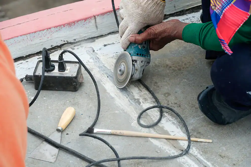 A person wearing gloves uses a handheld angle grinder to cut a groove in concrete. Nearby are a power strip, screwdriver, and paintbrush—essential tools often sourced from equipment rental St. Lucie & Orange County, FL.