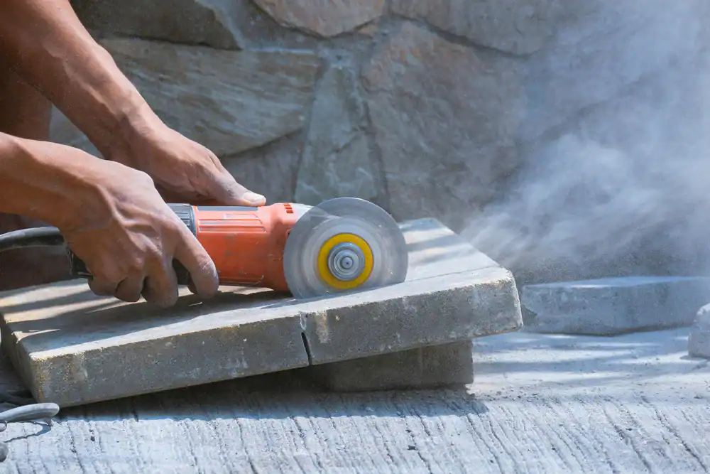 A person in FL uses an electric angle grinder, possibly from equipment rental St. Lucie & Orange County, to cut a concrete slab outdoors, sending dust clouds into the air as sunlight casts shadows and a stone wall stands in the background.