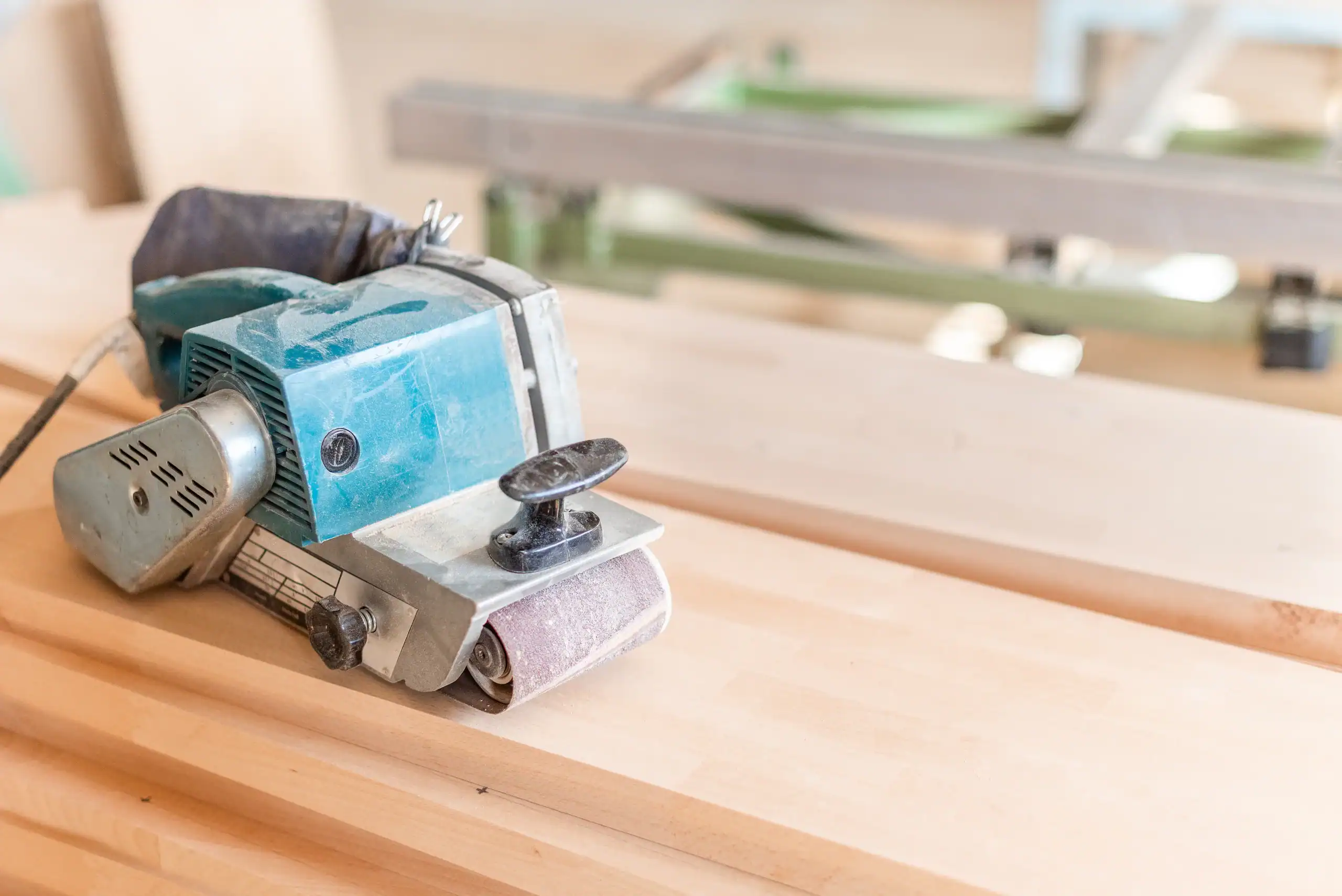 An electric hand sander rests on a wooden workbench in a bright workshop, with unfinished wood planks and blurred machinery visible&mdash;perfect for those seeking equipment rental in St. Lucie & Orange County, FL.