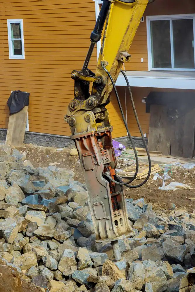 A hydraulic breaker attached to an excavator is breaking large rocks at a construction site in front of a yellow building—dust and debris swirl around this equipment rental St. Lucie & Orange County, FL relies on for tough jobs.