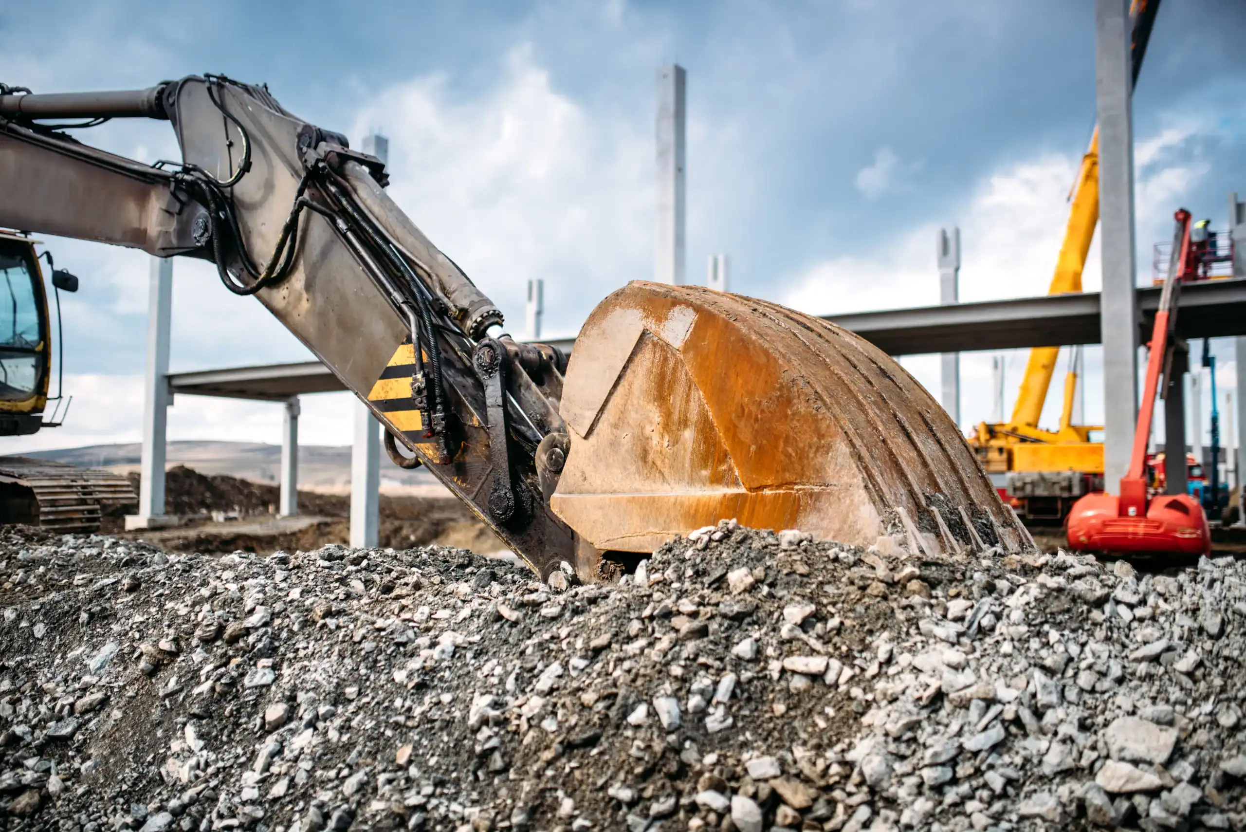 A close-up of an excavator&rsquo;s bucket digging into gravel at a construction site, with steel beams and equipment visible in the background under a cloudy sky&mdash;perfect for showcasing equipment rental St. Lucie & Orange County, FL.