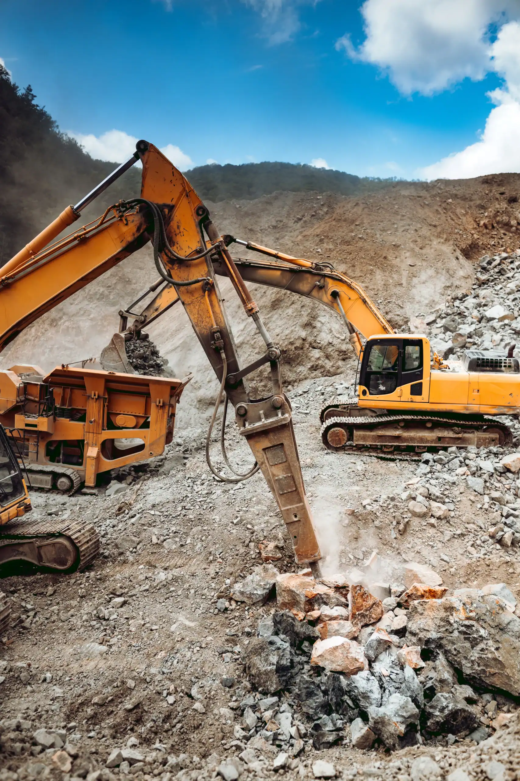 A yellow excavator with a hydraulic breaker attachment breaks rocks at a quarry site, surrounded by loose stones and heavy machinery&mdash;ideal for equipment rental St. Lucie & Orange County, FL&mdash;under a partly cloudy sky.