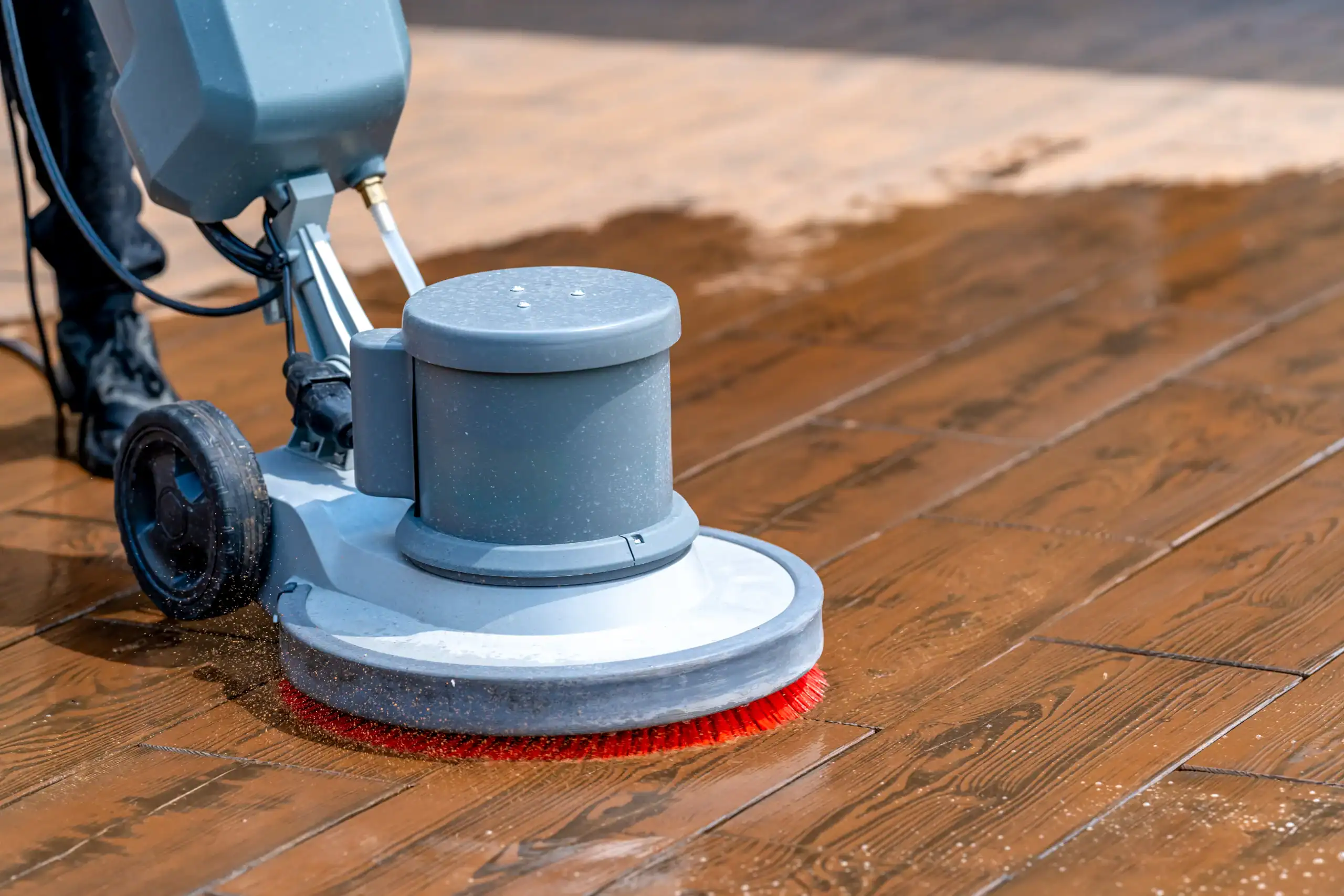 A floor cleaning machine with a red brush attachment is scrubbing a wooden floor, removing dirt and making the surface shine. Perfect for equipment rental St. Lucie & Orange County, FL.