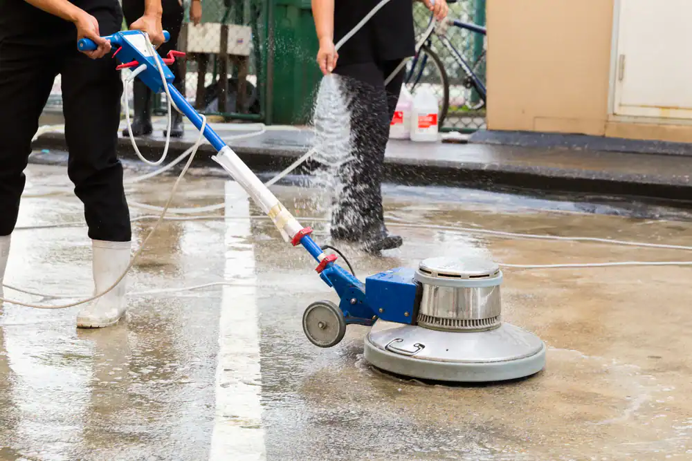A person in black clothing and white boots uses a floor cleaning machine from an equipment rental St. Lucie & Orange County on a wet concrete surface while another person sprays water nearby in FL.