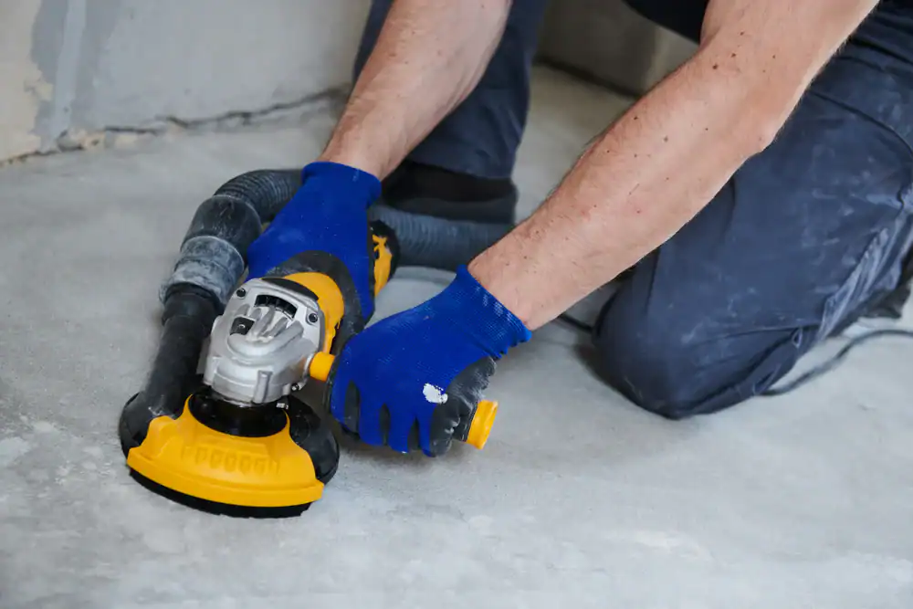 A person wearing blue gloves uses a yellow and black power sander, available through equipment rental St. Lucie & Orange County, FL, to smooth a concrete floor with a vacuum attachment connected for dust collection.