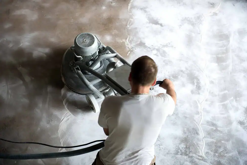 A person in a white t-shirt operates a floor polishing machine on a dusty concrete surface, smoothing and cleaning the floor—equipment rental St. Lucie & Orange County, FL makes this task easier. The image is taken from above and behind.