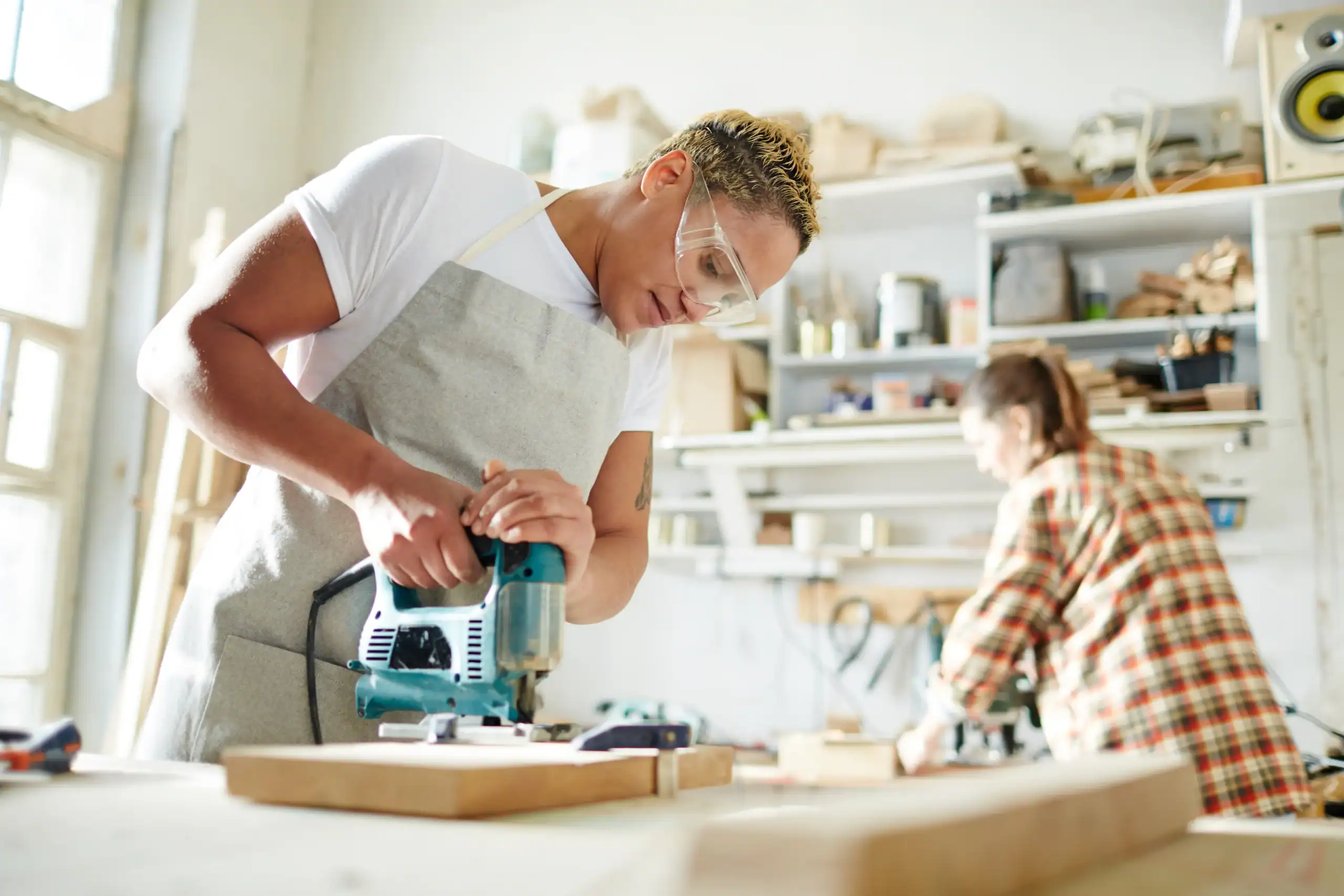 A person wearing safety goggles and an apron uses a jigsaw to cut wood in a workshop, showcasing the benefits of equipment rental St. Lucie & Orange County, FL. Another person works at a bench, surrounded by tools and shelves.
