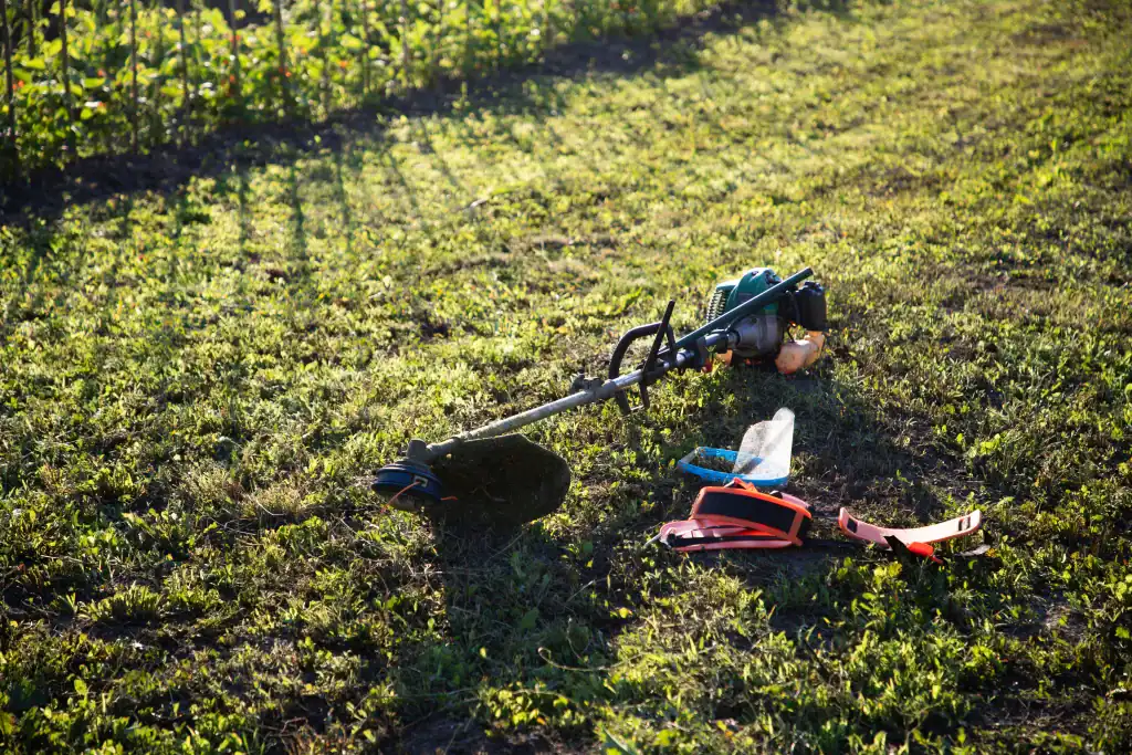 A grass trimmer and safety gear, including gloves and a face shield, lie on a sunny lawn in FL, with a garden fence and green plants in the background—ideal equipment rental for St. Lucie & Orange County projects.