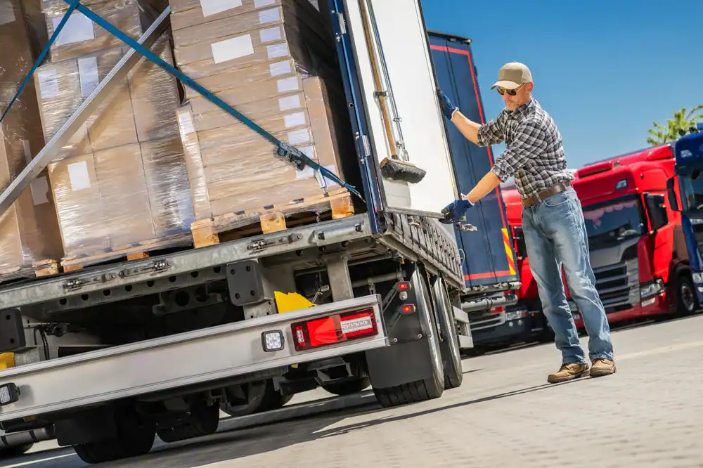 A man in a plaid shirt and cap stands beside a truck, closing its rear door. Pallets of boxes are visible inside, with FL equipment rental St. Lucie & Orange County trucks parked nearby under a clear blue sky.