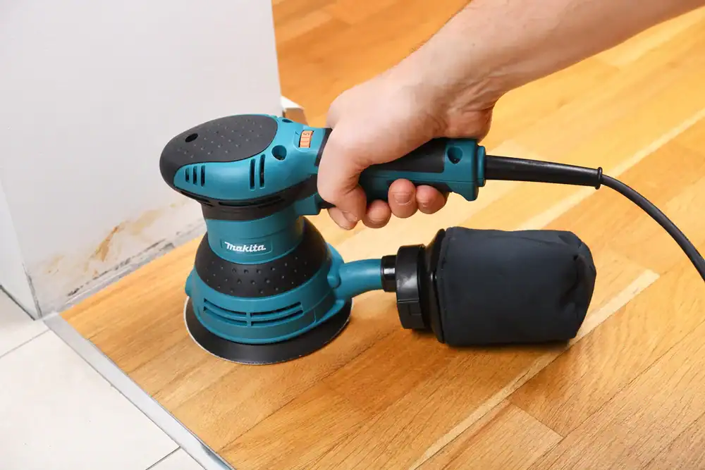 A person uses an electric sander with a dust bag, available through equipment rental St. Lucie & Orange County, FL, to smooth the surface of a wooden floor near a wall and white tile.