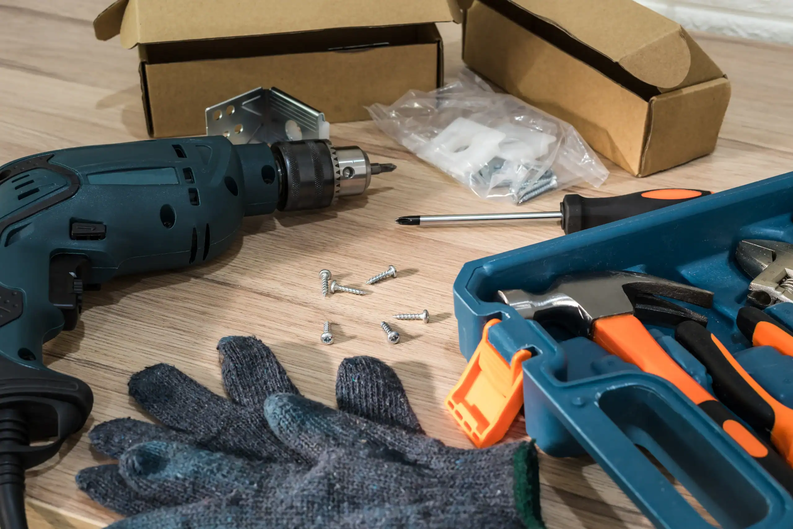 A close-up of various tools on a wooden floor, including a power drill, screwdriver, hammer, pliers, and work gloves&mdash;perfect for equipment rental St. Lucie & Orange County, FL&mdash;with cardboard boxes and hardware in the background.