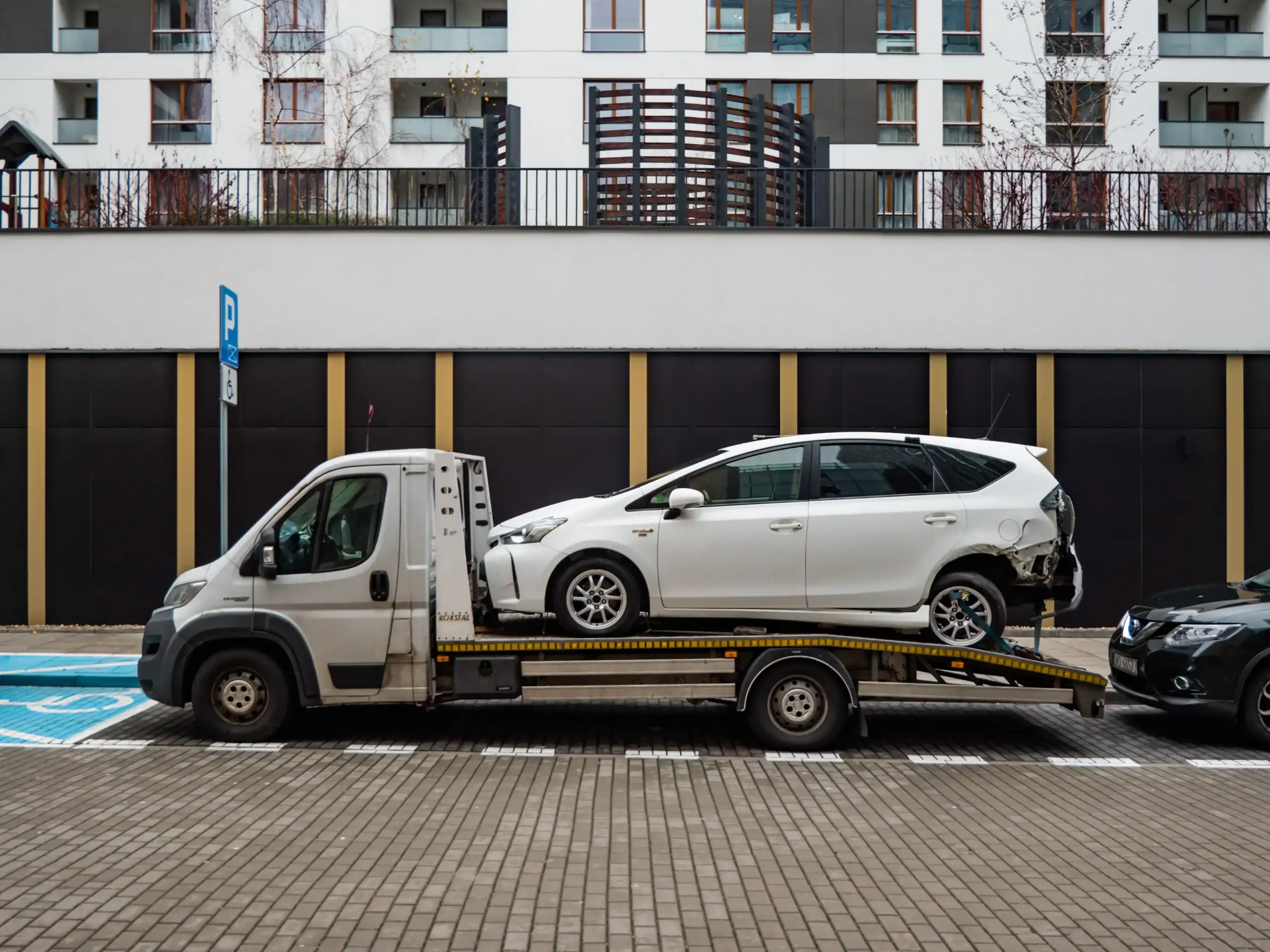 A tow truck carrying a damaged white car is parked on a city street in FL, in front of modern apartment buildings. The car appears to have rear-end damage, possibly awaiting equipment rental St. Lucie & Orange County services.