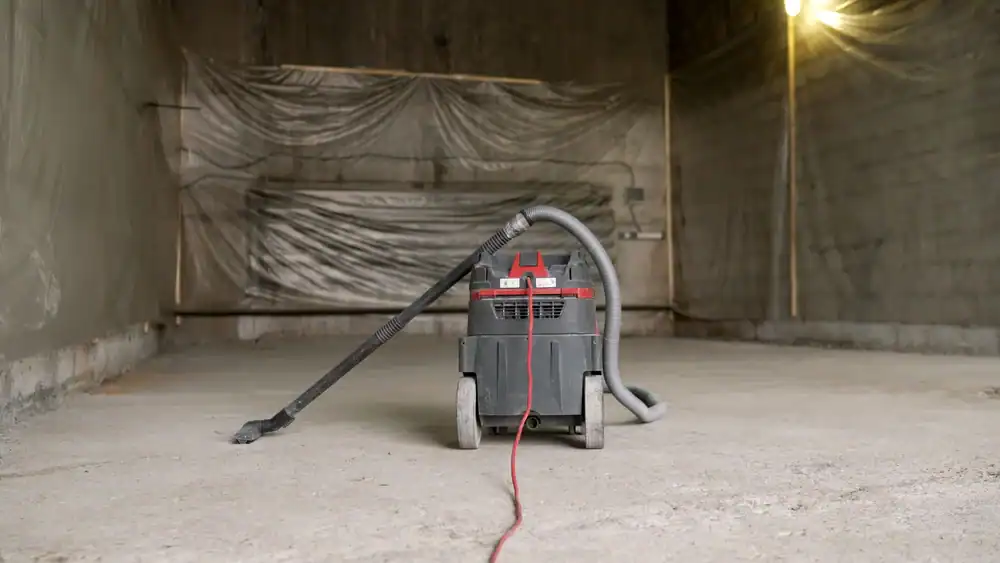 A shop vacuum stands in the middle of an unfinished, empty room with concrete floors and plastic sheeting covering the walls—showcasing equipment rental St. Lucie & Orange County, FL under a single glowing bulb.
