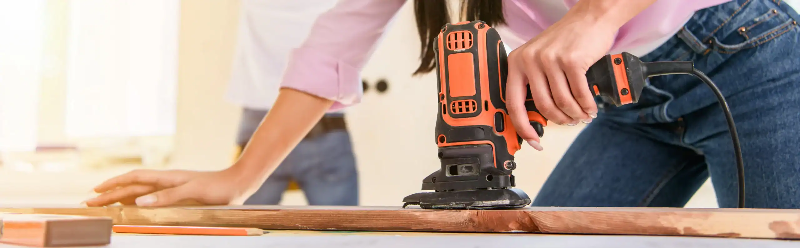Close-up of a person using an electric sander, likely sourced from equipment rental St. Lucie & Orange County, FL, to smooth a wooden plank. Another person is blurred in the background, while the one sanding wears a light pink shirt and jeans.
