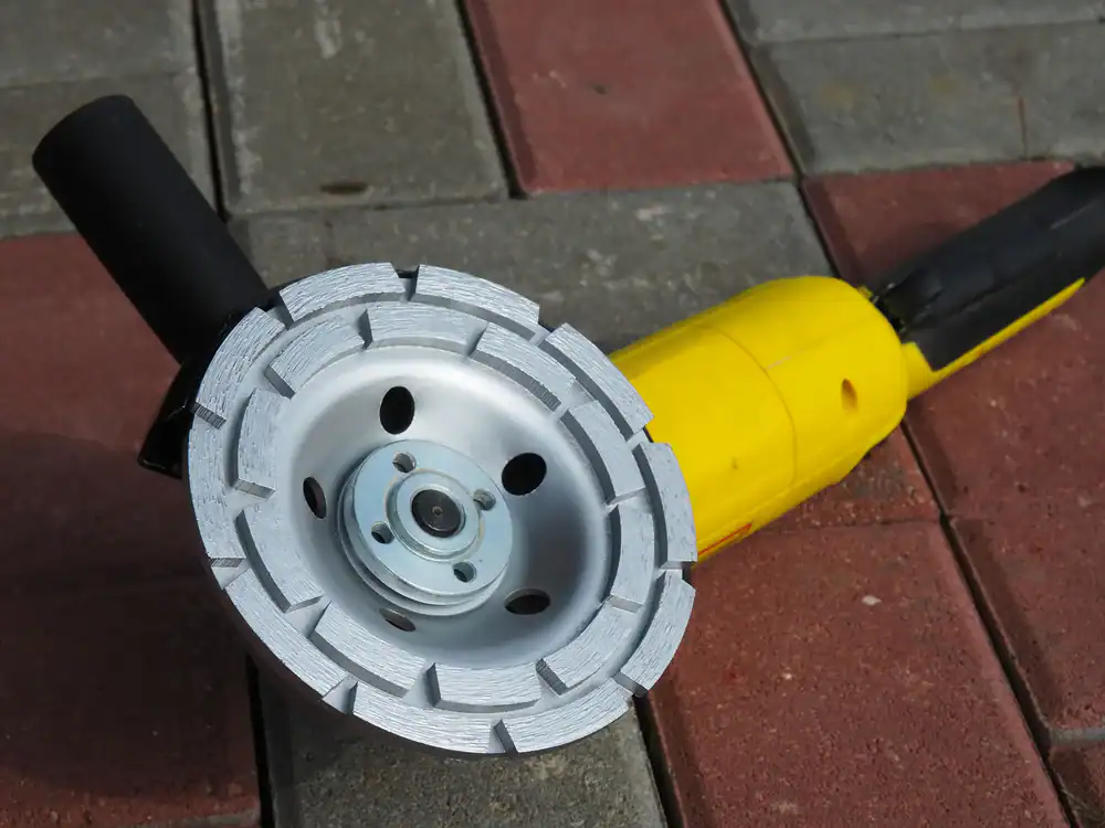 A yellow handheld angle grinder with a silver diamond blade rests on red and gray paving stones outdoors. This durable FL equipment rental St. Lucie & Orange County tool features a black handle for easy use.