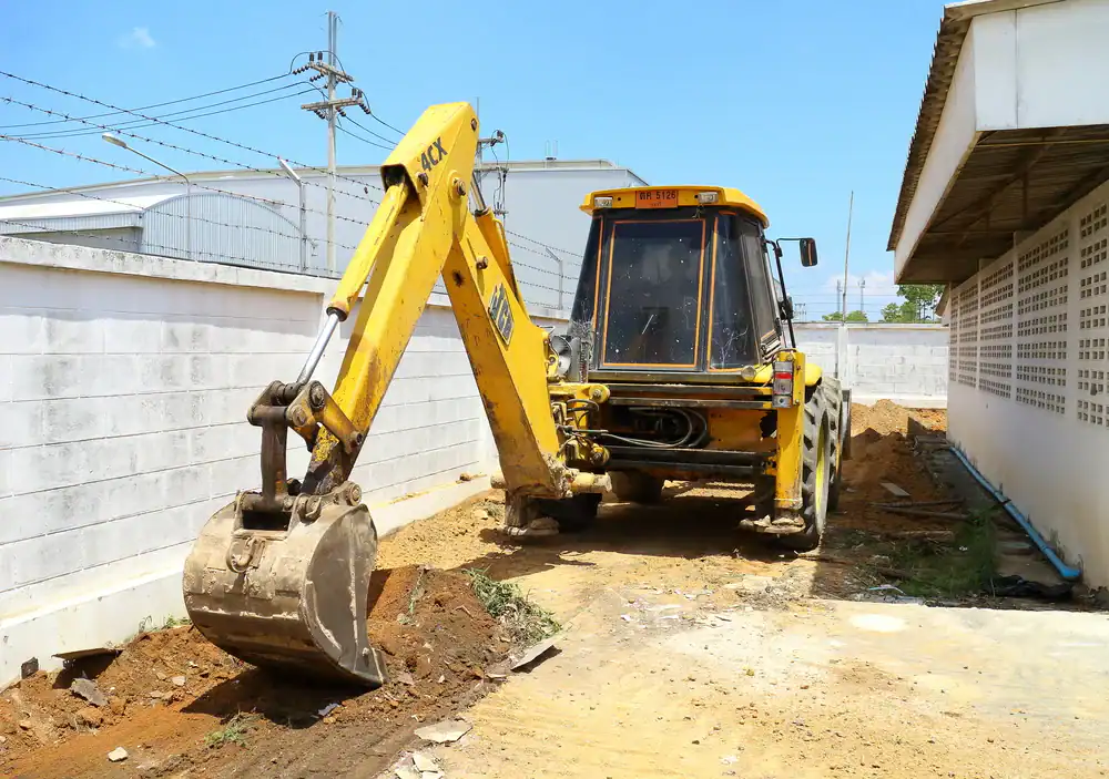 A yellow backhoe loader from an equipment rental St. Lucie & Orange County is digging a narrow trench next to a white concrete wall at an outdoor construction site under the clear blue FL sky.