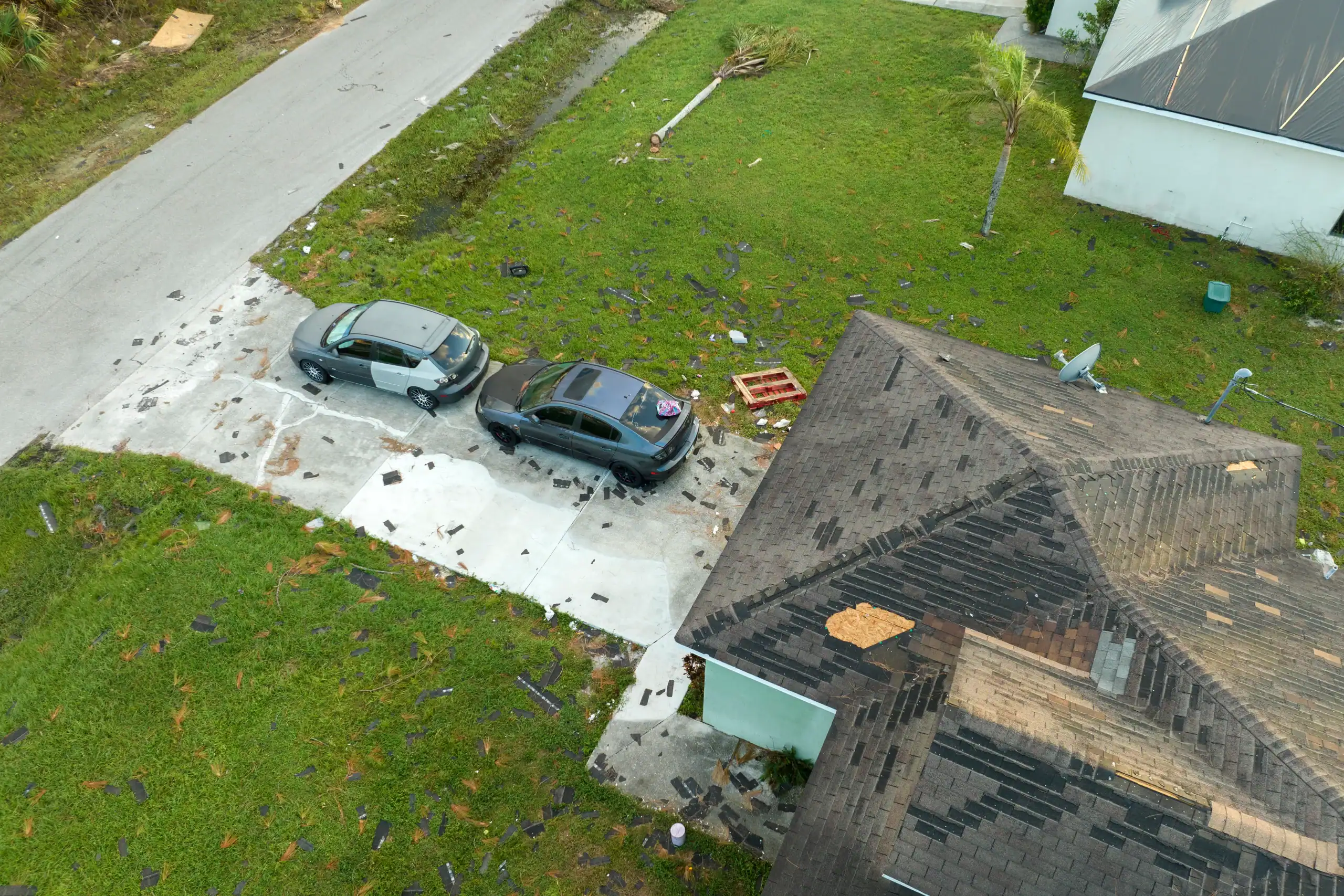 Aerial view of two cars parked in a driveway with scattered roof shingles and debris on the ground and lawn, next to a house with visible roof damage.