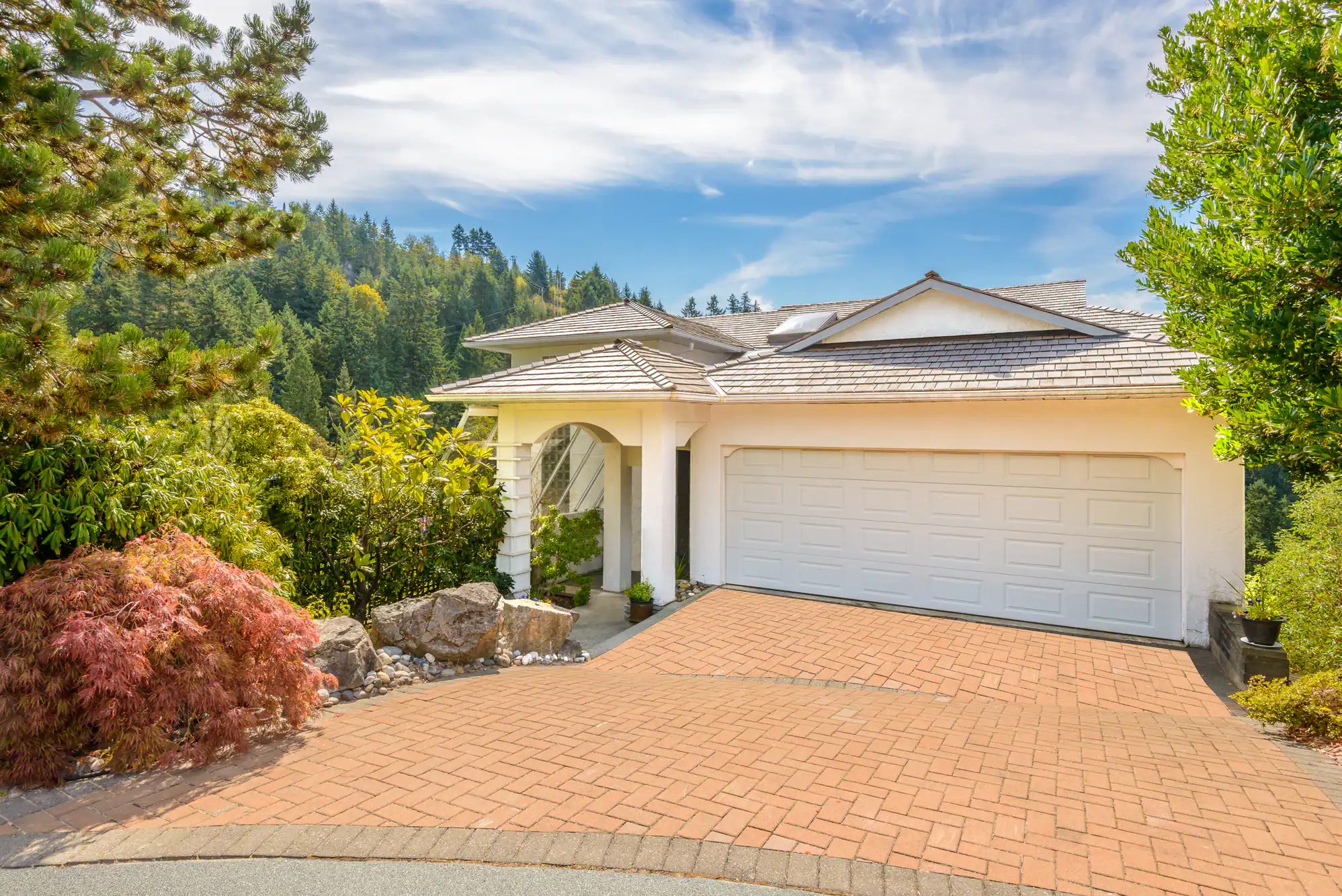 A modern white house with a two-car garage and tiled roof sits on a brick driveway, surrounded by lush trees and greenery, under a bright blue sky with scattered clouds.
